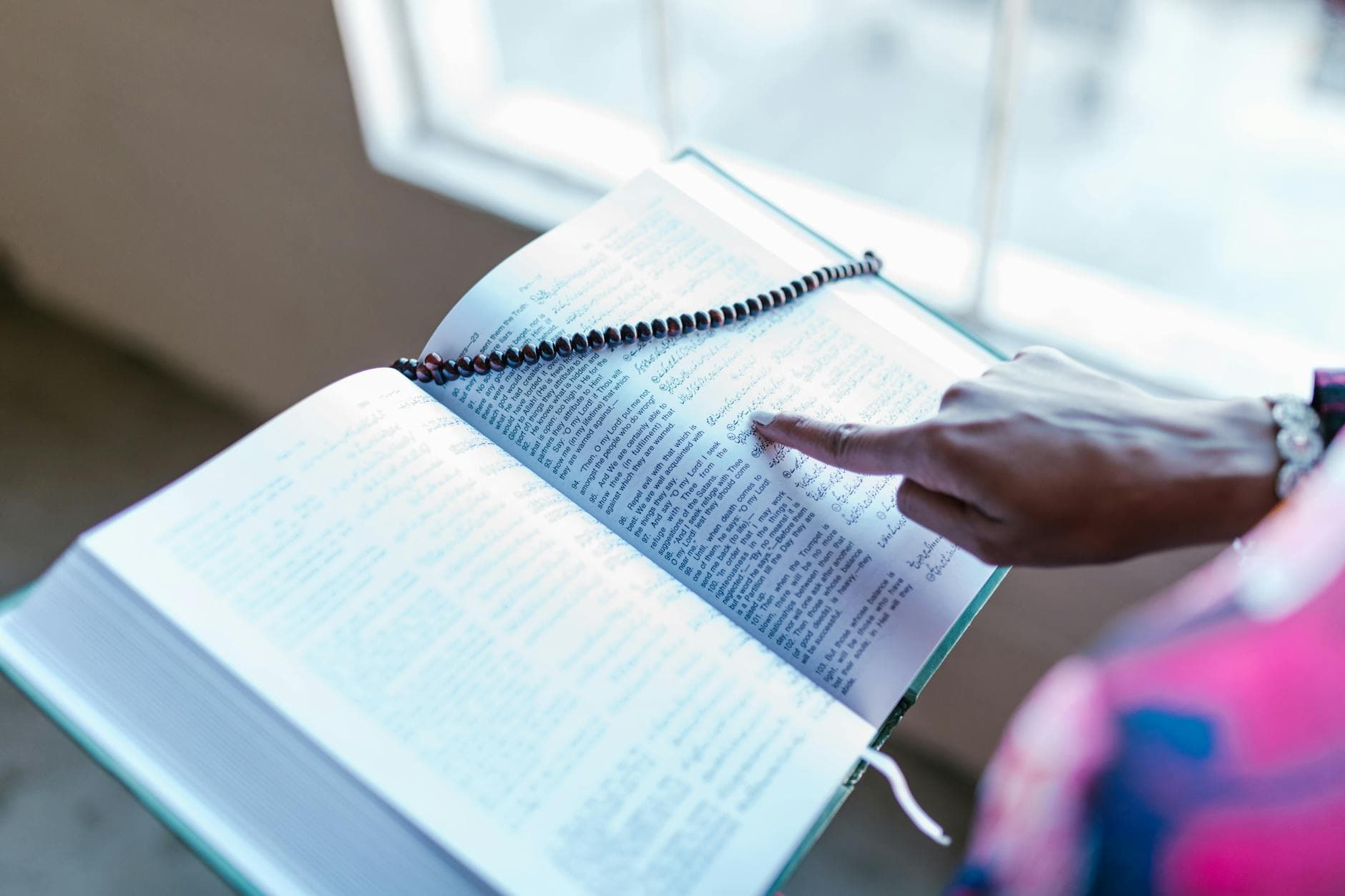 Photo by RDNE Stock project on Pexels A Muslim woman pointing to a passage in the Quran with prayer beads draped over it indoors.