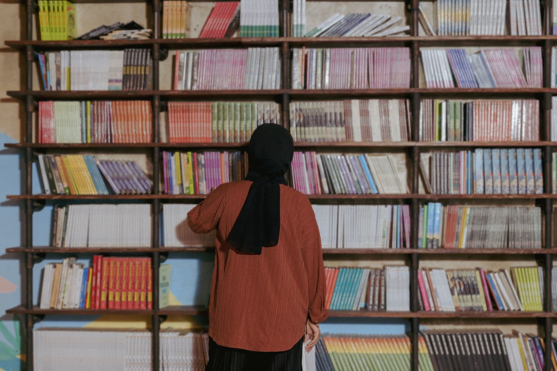 Photo by Yazid N on Pexels A woman in a headscarf selects a book from a library filled with colorful volumes.